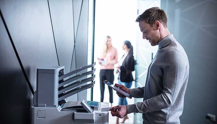 Férfi, aki NFC-kártyát tart a nyomtatóhoz, tablet a kezében, emberek a háttérben