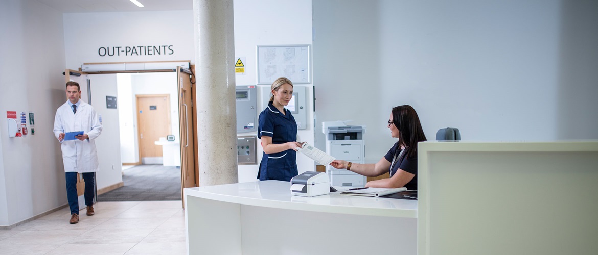 Szemüveget viselő recepciós hölgy ül az asztalnál, a háttérben egy nővér és egy orvos dolgoznak