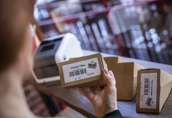 Egy nő ellenőrzi a Brother címkézőgép nyomtatott élelmiszer-címkéit, amelyeket egy élelmiszercsemege vagy pékség boltban előrecsomagolt szendvicsekre ragasztottak. A címkék vonalkódot, címet, képet, árat és dátum szerinti felhasználást tartalmaznak.