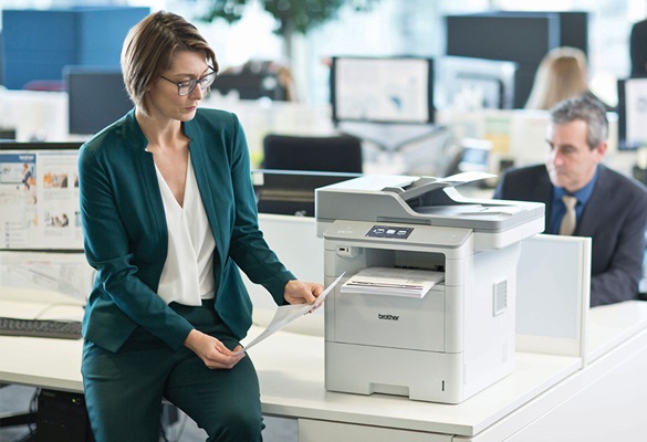 Szemüveges hölgy zöld kosztümöt visel és ül az asztalon egy Brother MFC-L6900DW nyomtató mellett, öltönyös férfi, monitor, asztal a háttérben